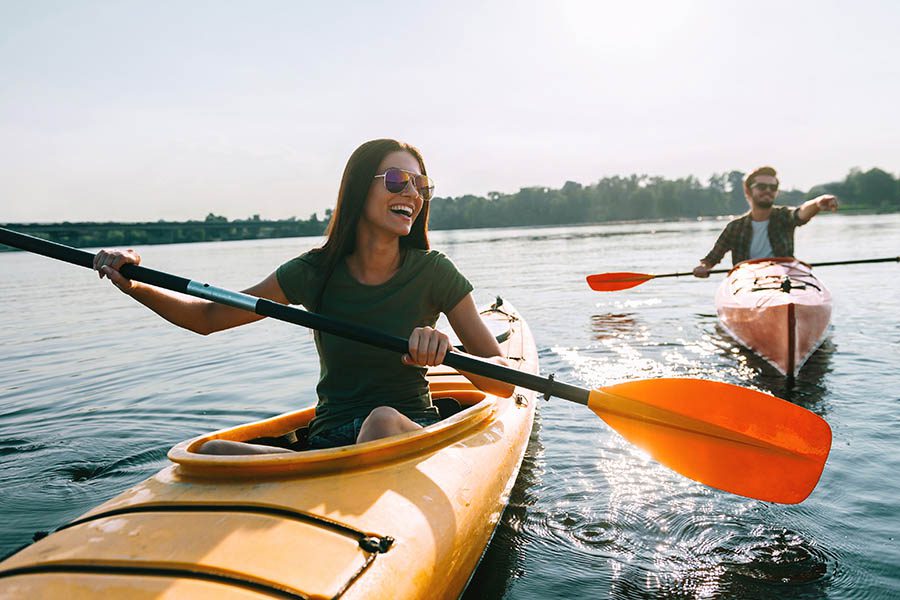 About Our Agency - Young Couple Paddle Single Kayaks on a Tree-Lined Lake, Smiling and Pointing up Ahead