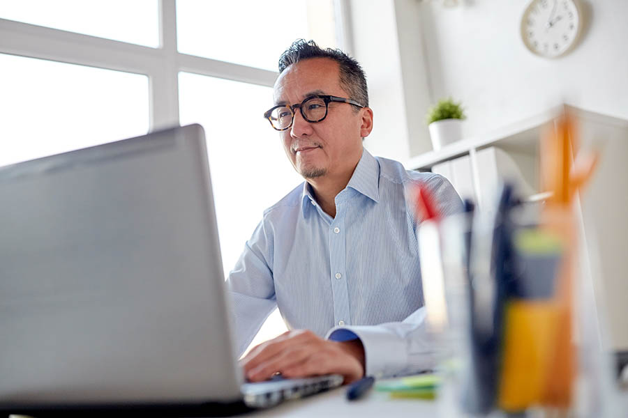 Blog - Middle-Aged Man in Dress Shirt and Tortoiseshell Glasses Reads on His Laptop in a Modern, Clean White Office