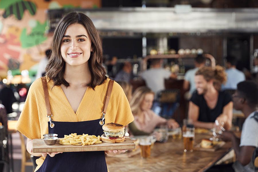 Business Insurance - Young Waitress Holds a Cheeseburger Meal on a Wooden Plank, as Customers Chat and Drink Beer in the Bustling Restaurant Behind Her