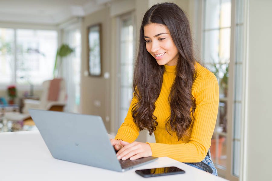 Account Management - Young Woman in Yellow Turtleneck Sweater Uses a Laptop at a White Countertop in Her Bright Apartment