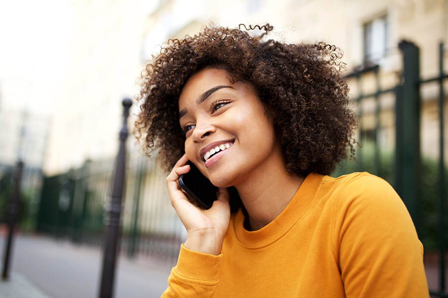 Contact Us - Smiling Black Woman in Yellow Sweater Sits Outside Making a Call on Her Cell Phone