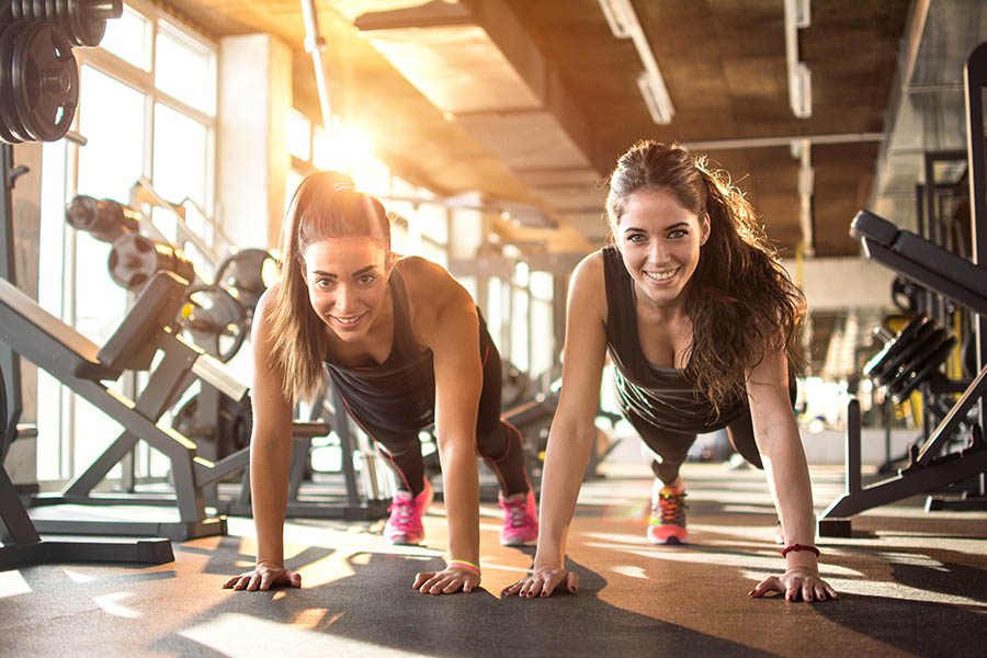 Employee Benefits - Two Smiling Female Friends Do Pushups Side by Side on the Floor of a Gym
