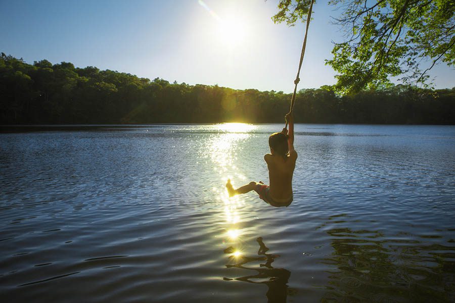 Lake Villa, IL Insurance - Young Boy in a Swimsuit Swings on a Rope Swing From a Tree Branch Into a Lake With the Sun Reflecting off the Water